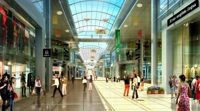 Modern shopping mall corridor with glass ceiling, stores on both sides, and shoppers walking.