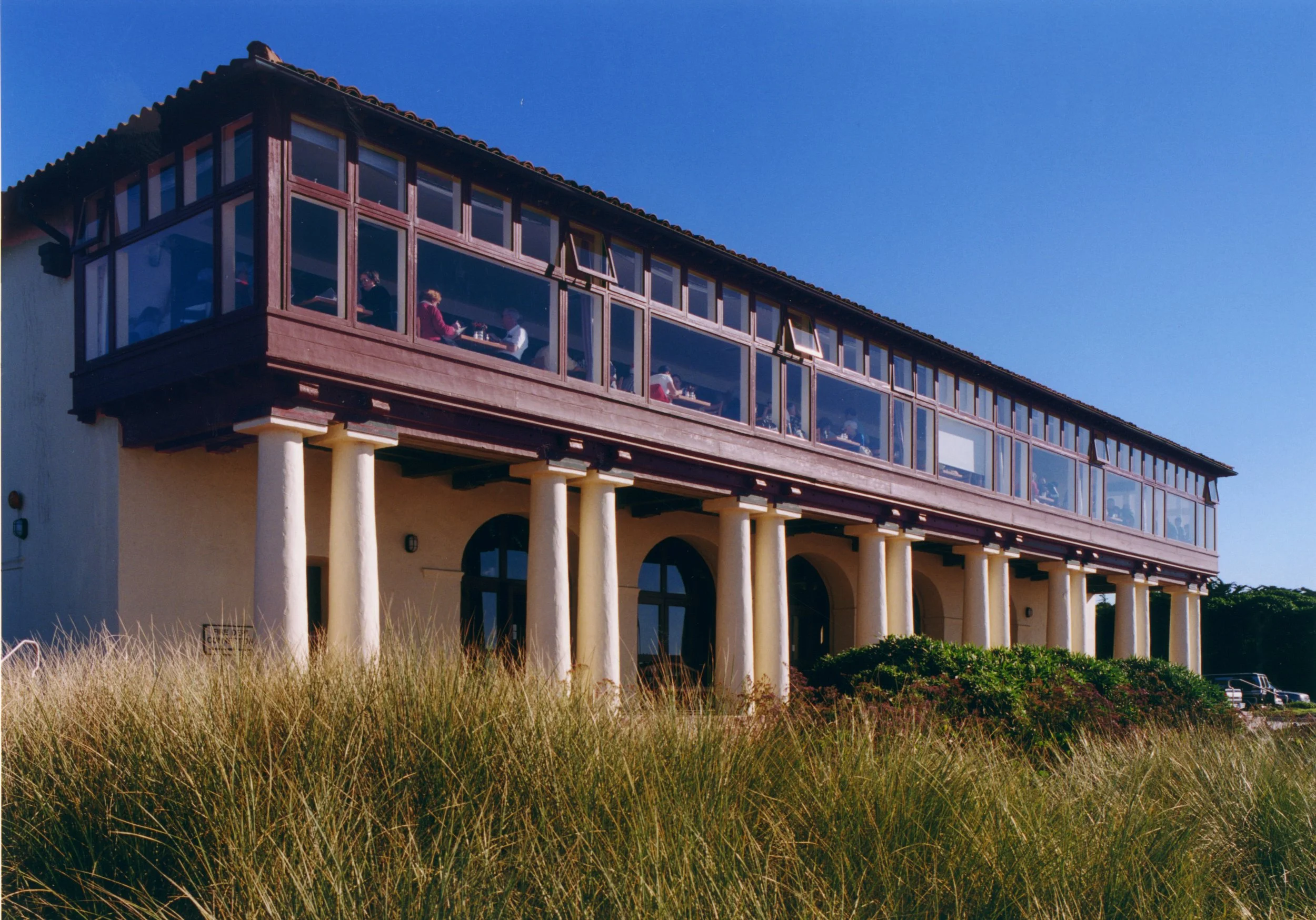A large building with an upper level enclosed in glass windows and a lower level supported by white columns, situated in a natural landscape with tall grass, under a clear blue sky.