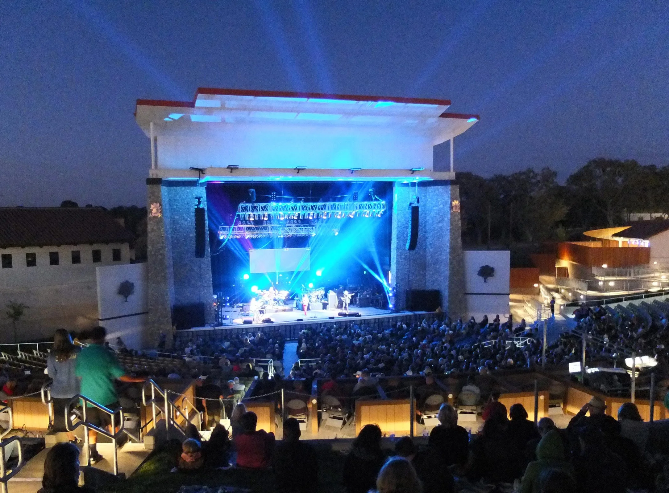 An outdoor theater with a live music concert happening at night, audience seated in front of the stage, which is illuminated with blue and purple lights.