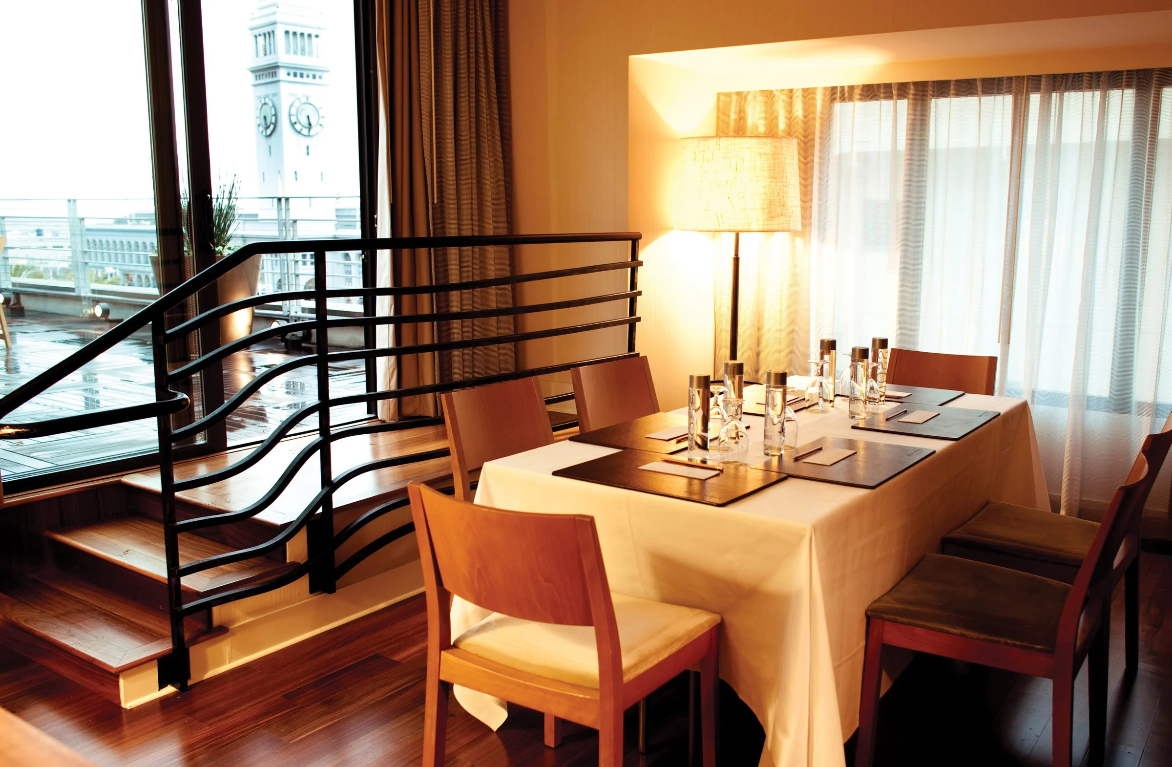 Elegant restaurant dining table set with placemats, napkins, glasses, and water bottles, near large windows with curtains, a lamp, and a staircase, overlooking a clock tower outside.