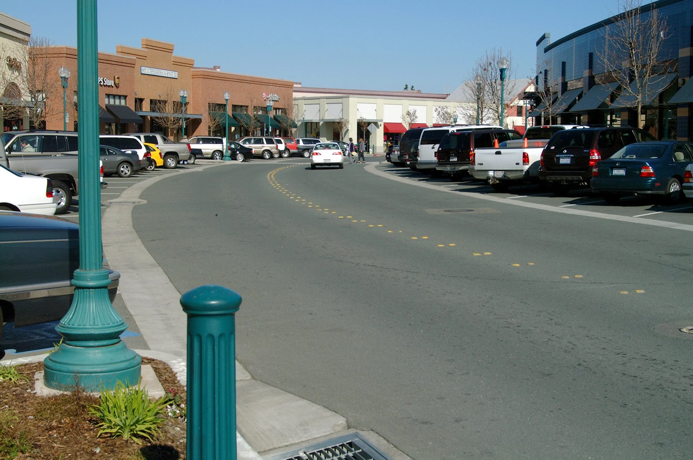 A parking lot outside a shopping center with cars parked along the curb and pedestrians walking, with storefronts and stores in the background under a clear blue sky.