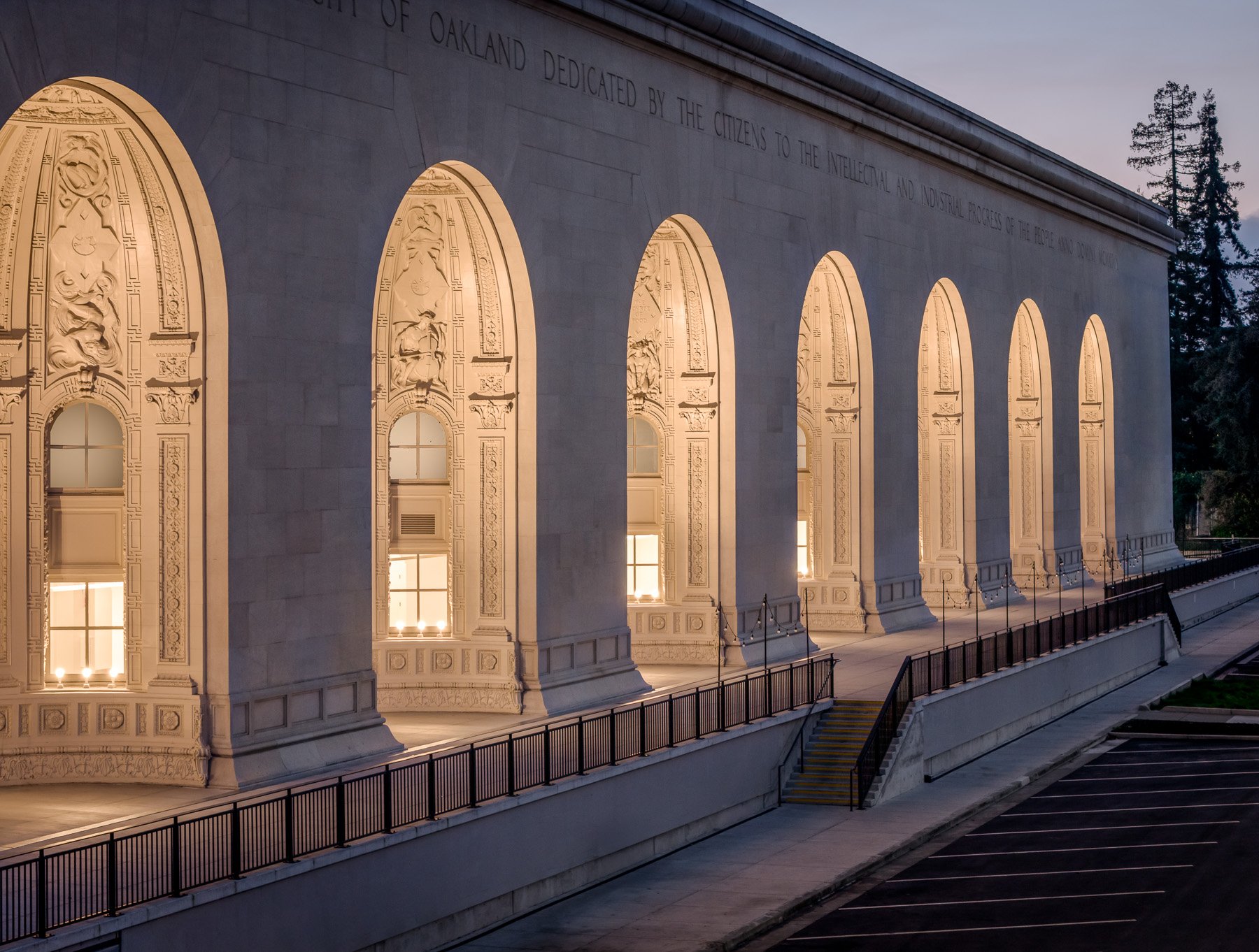 Side view of a large stone building with illuminated arched windows and intricate decorative reliefs, surrounded by a parking lot and trees in the background at dusk.