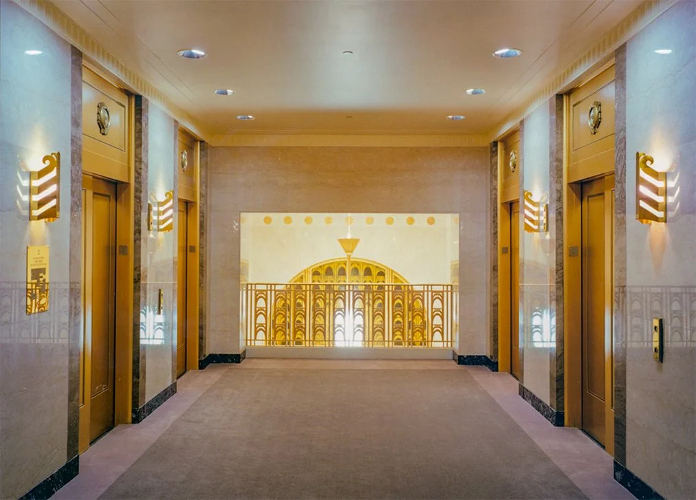 The image shows a hallway with four golden elevator doors, two on each side. The hallway features a decorative wall with a large artwork or mural at the end, including a golden dome structure and symmetrical patterns. The ceiling has recessed lightin