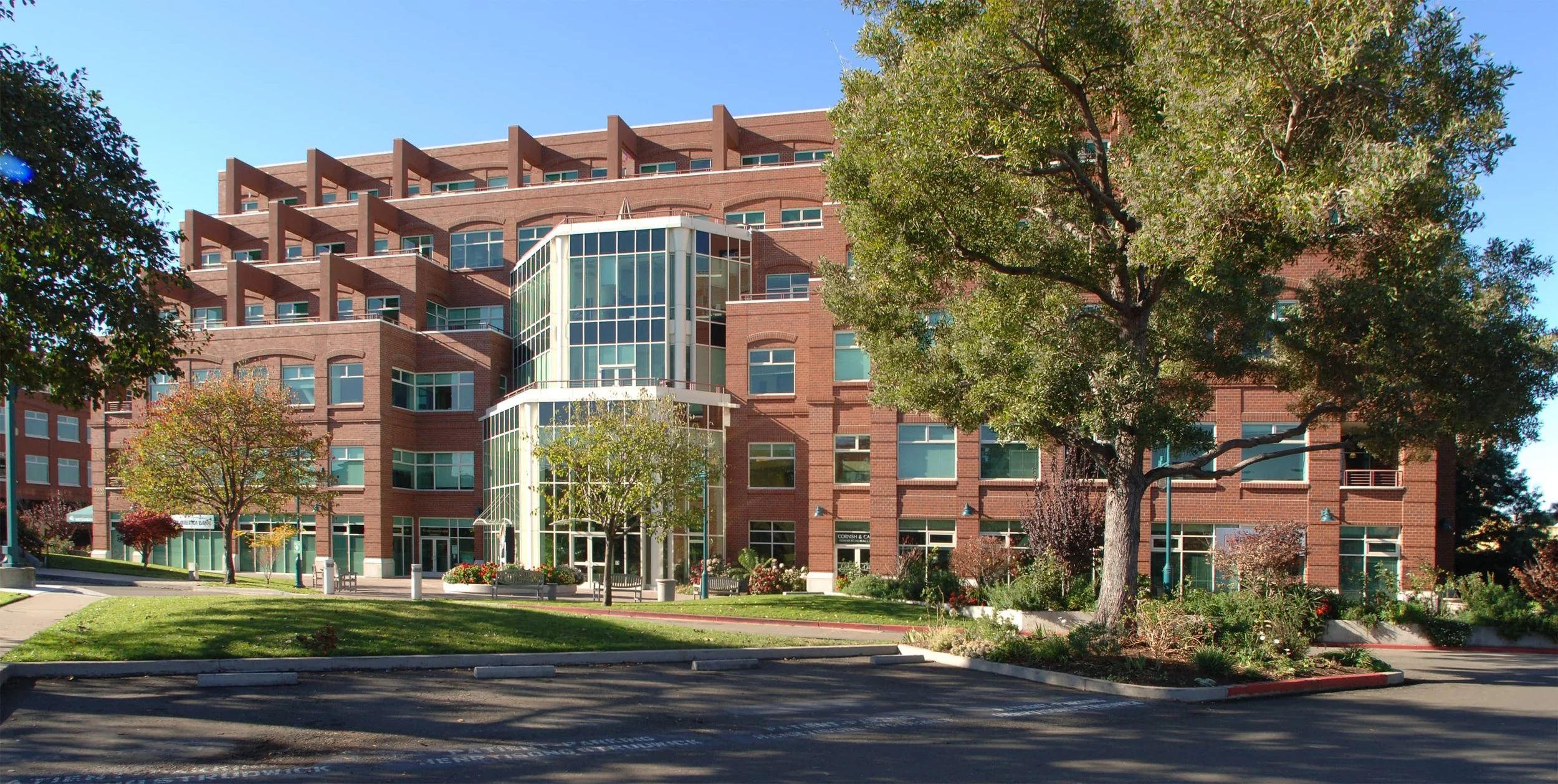 A modern multi-story office building with a brick facade and green-tinted glass windows, surrounded by trees and landscaped greenery under a clear blue sky.