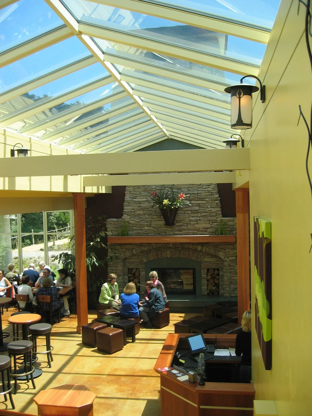 Interior of a sunny restaurant or cafe with a stone fireplace, wooden furniture, and a high glass ceiling, where people are seated and engaged in conversations.