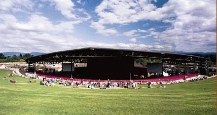 A large outdoor stadium with a covered seating area, set in a grassy field under a mostly cloudy sky.