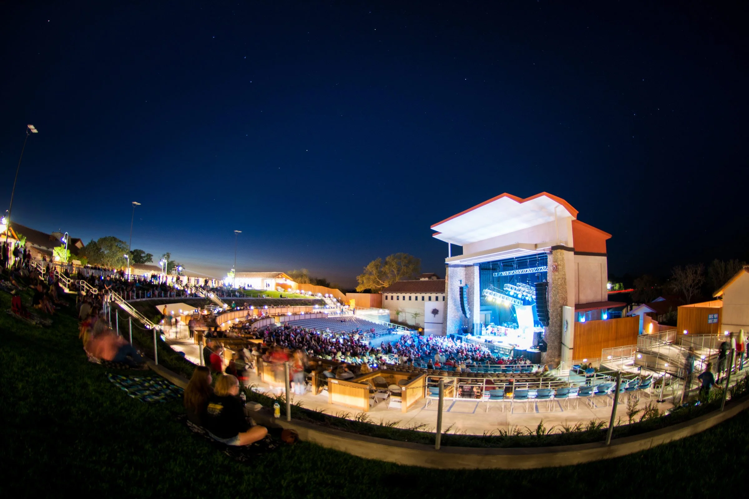 Nighttime outdoor concert at an amphitheater with a large crowd, stage lighting, and a starry sky.