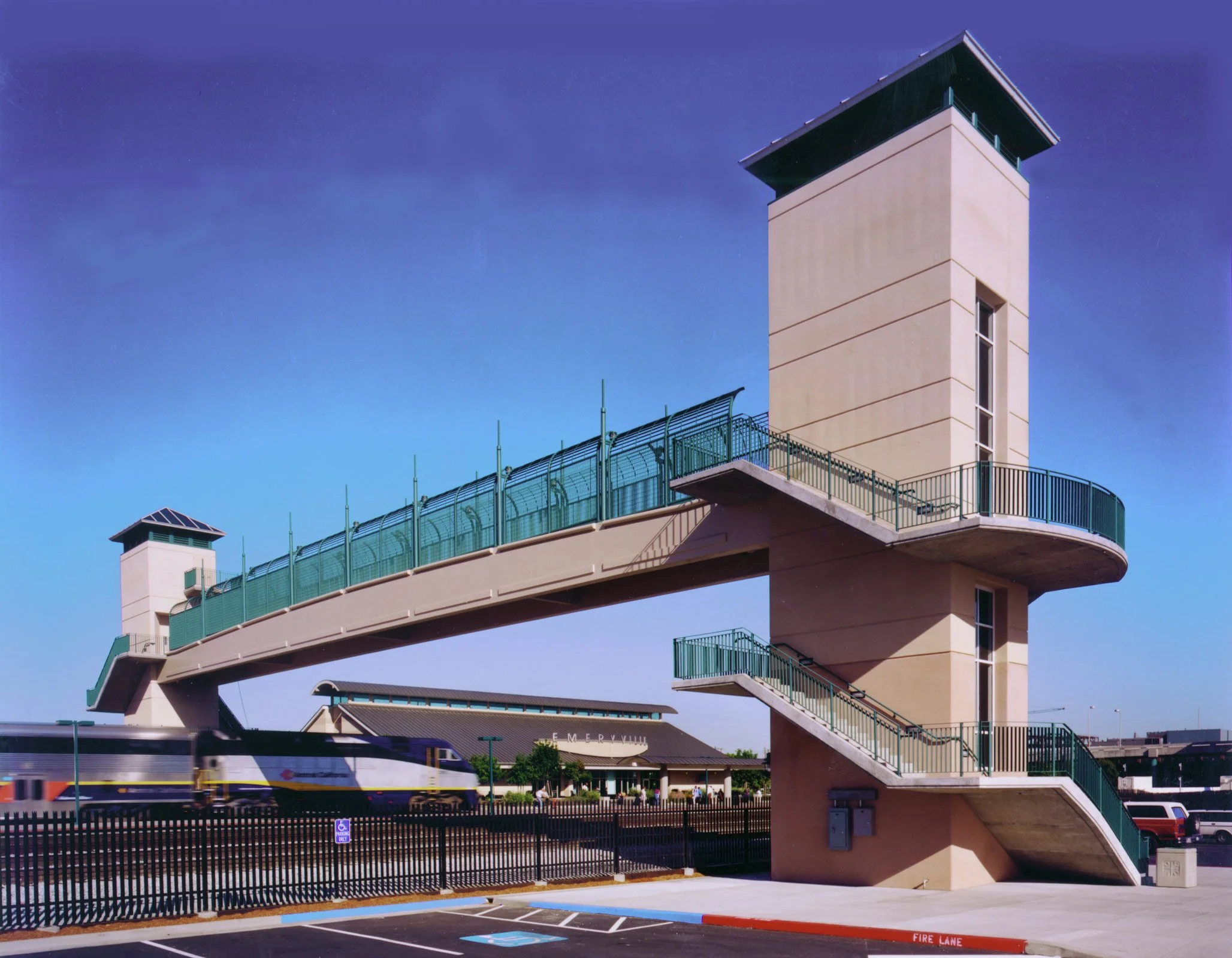 The image shows a bridge with staircases and tower structures, likely at a train station. A moving train is visible on the tracks below, and there is a building labeled "EMERYVILLE" in the background. The sky is clear and blue.