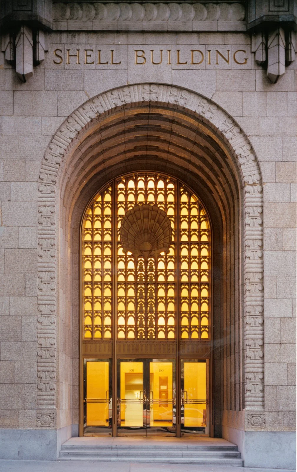 The entrance to the Shell Building features an ornate archway with intricate carvings and a large glass door, which has a grid of illuminated windows above it, framed by stone walls.