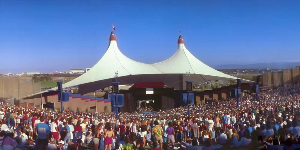 Large outdoor music concert with a big crowd gathered in front of a stage with a tent-like canopy.
