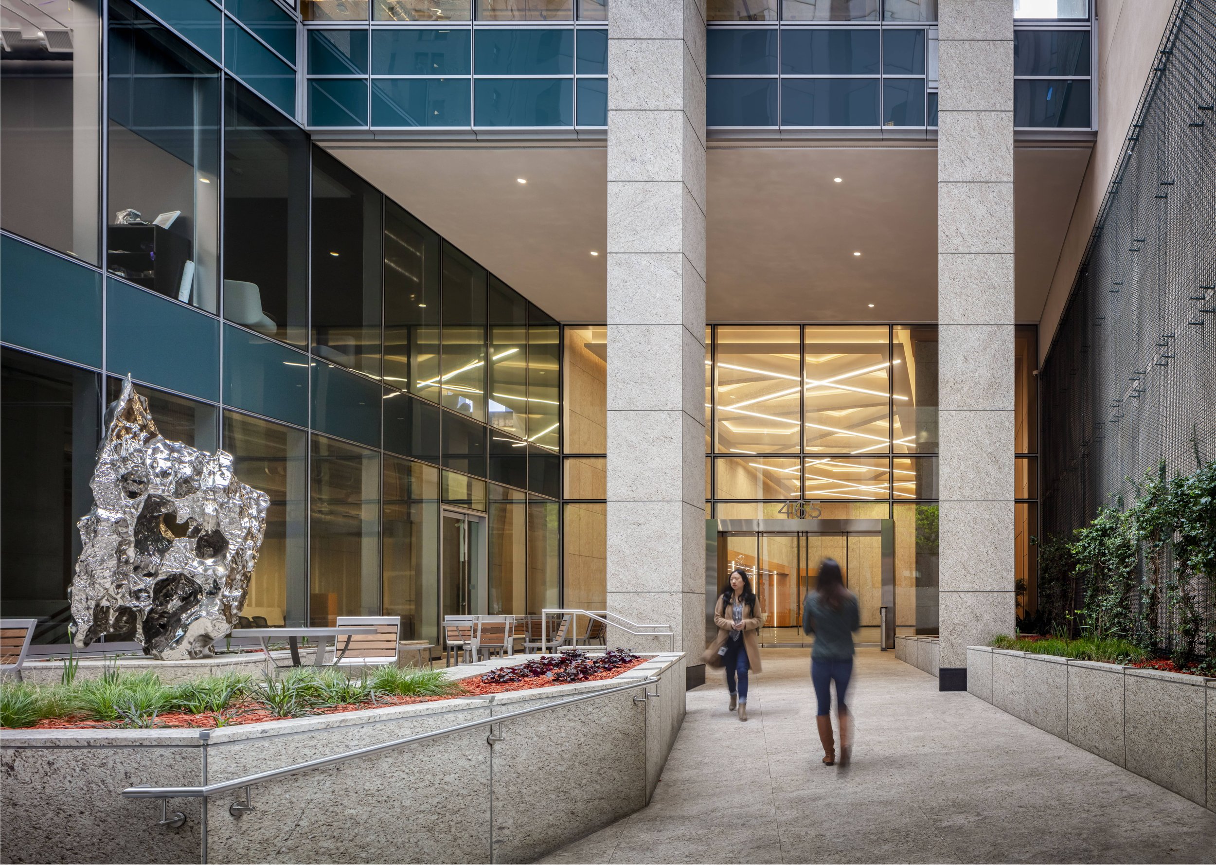 Modern office building entrance with glass windows/reflections, sculptures, benches, and two women walking in the courtyard.