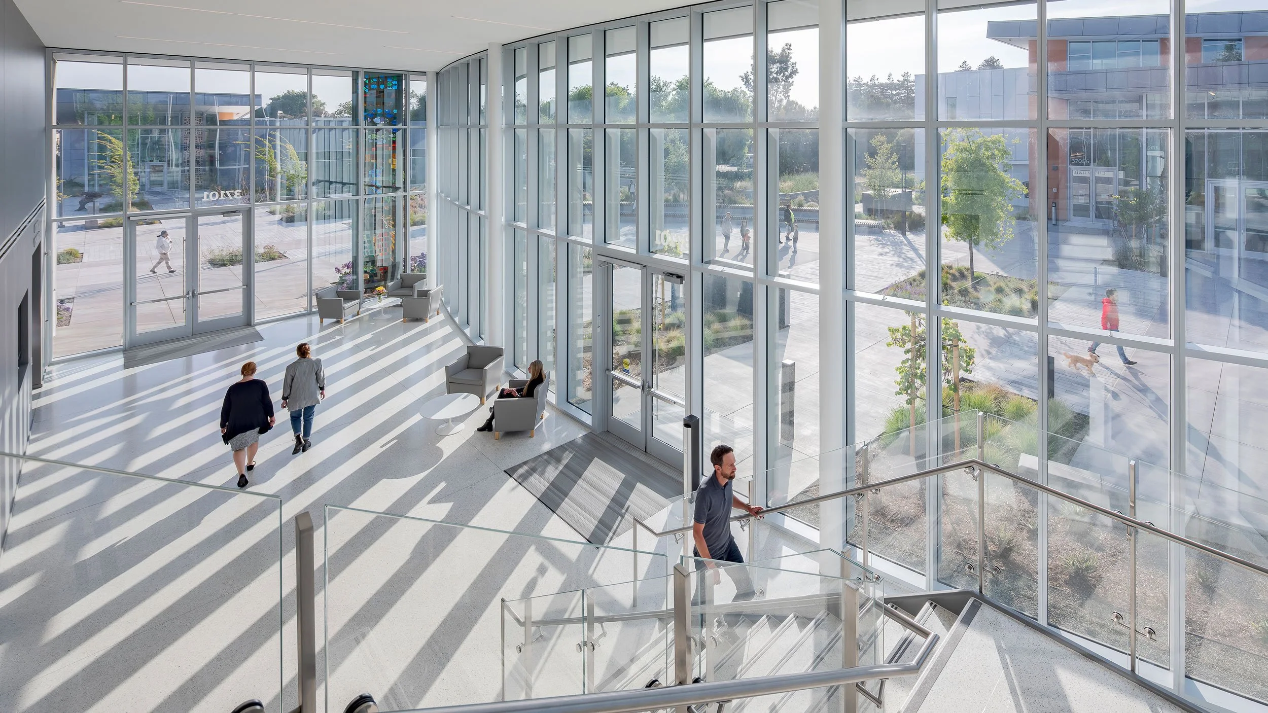 Bright, modern building lobby with large glass windows, sunlight casting striped shadows on the floor, people walking and sitting, and outdoor scene visible through windows.