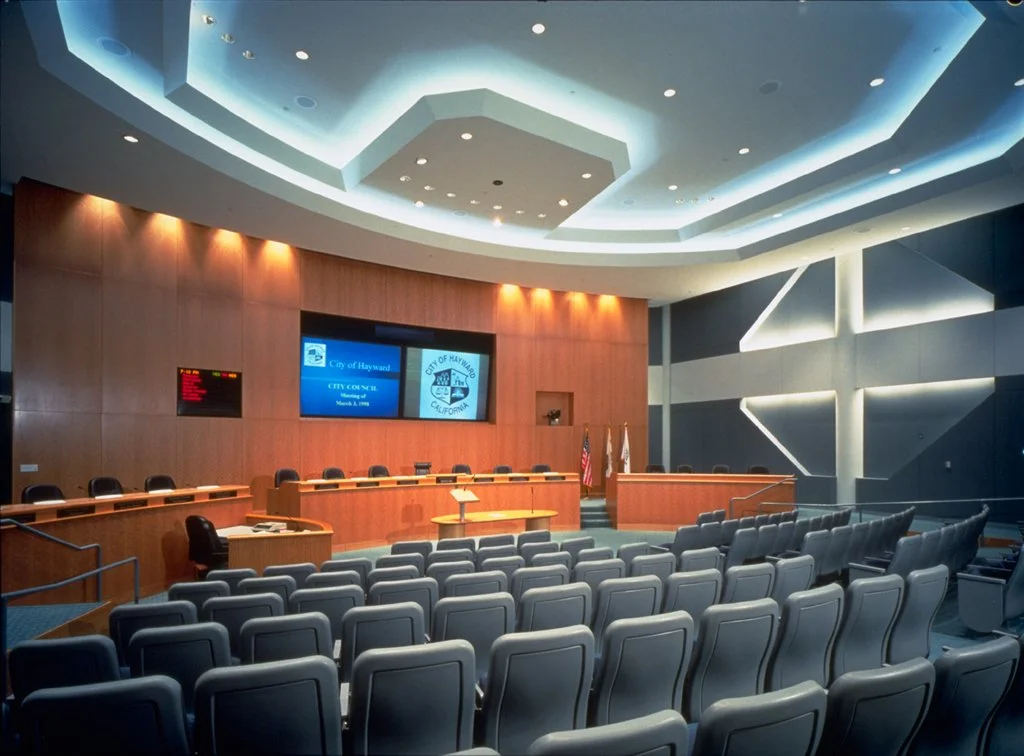 A modern city council meeting room with a curved wooden dais, multiple chairs, and large screens displaying the City of Hayward, California. The room has tiered seating and is illuminated with recessed lighting.