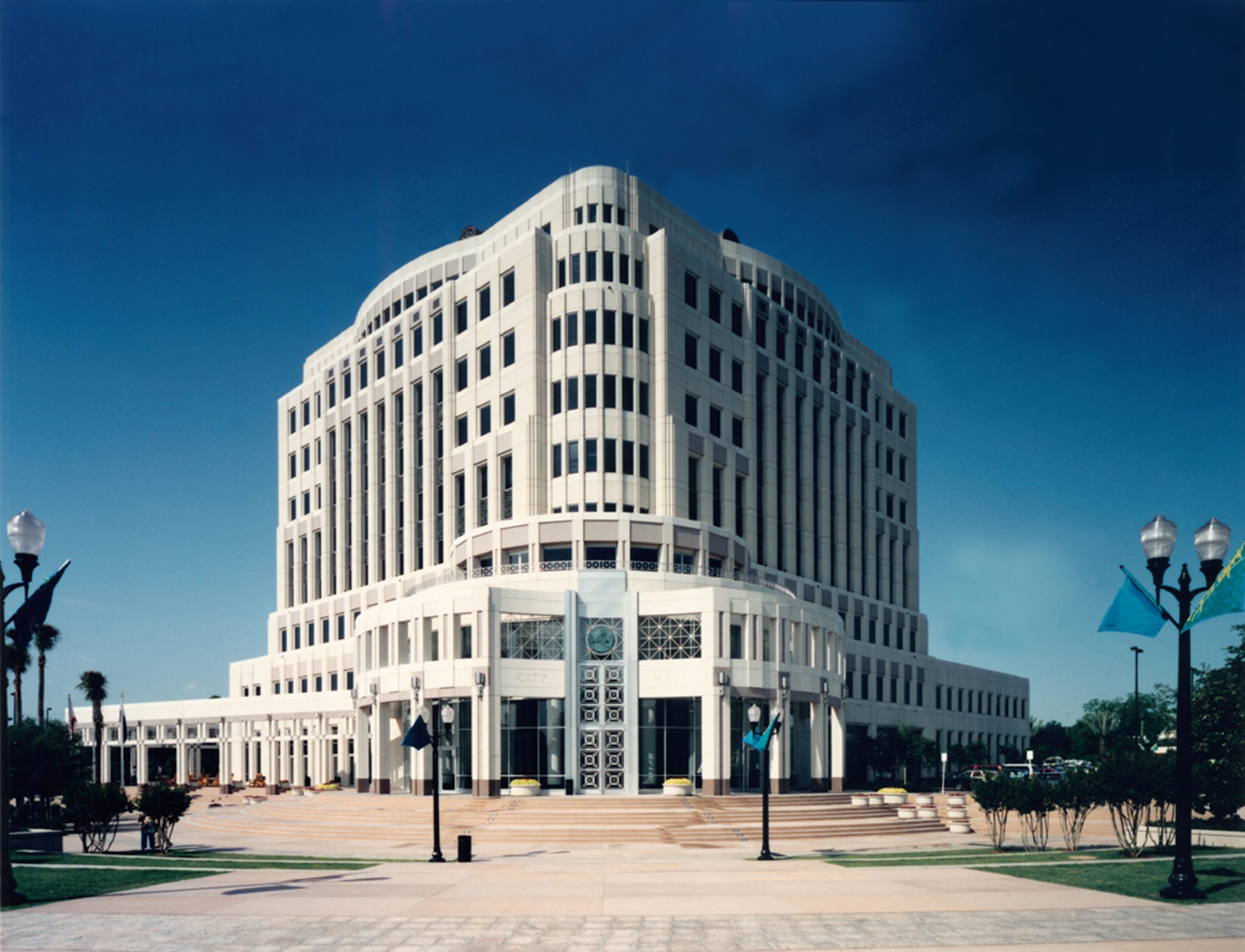 A tall modern white building with multiple windows, set against a clear blue sky, with a plaza and lampposts in the foreground.