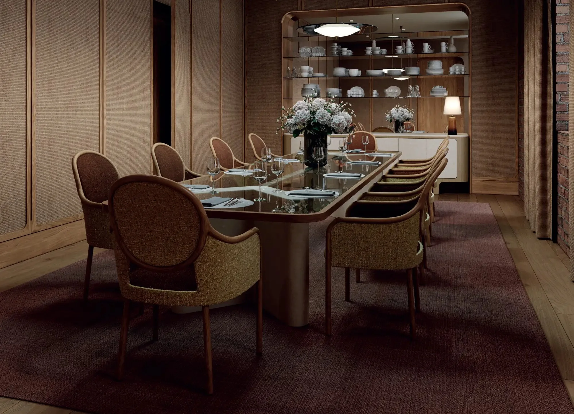 A dining room with a long table set with glasses, plates, and napkins. There are floral centerpieces and chairs arranged around the table. The room has warm lighting, wooden panel walls, and decorative shelves with dishware.
