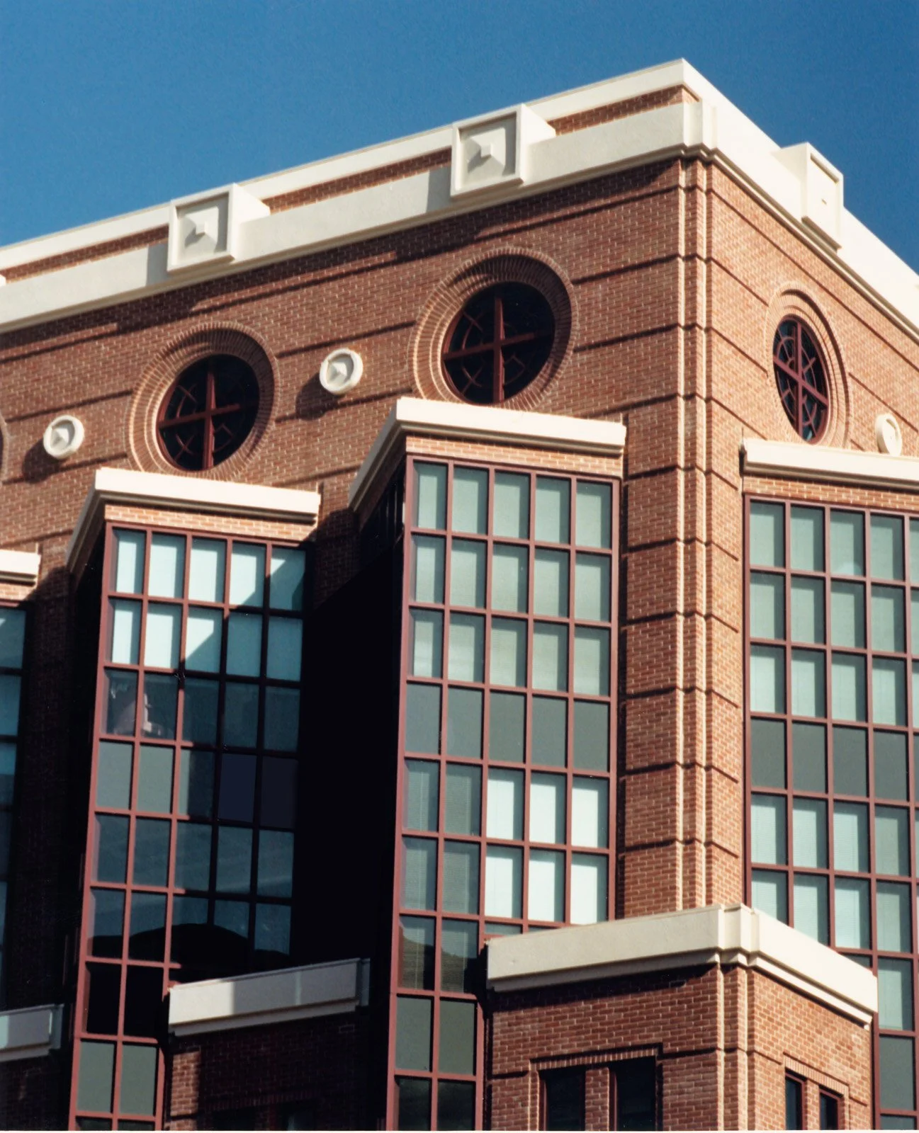 Close-up of a brick building with large glass windows and circular windows with red frames against a blue sky.