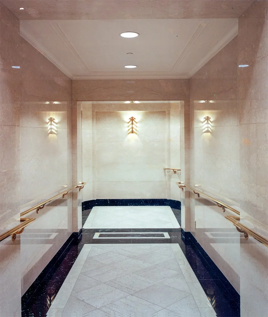 An elevator interior with beige marble walls, gold handrails, and wall-mounted light fixtures, reflecting in the mirrored walls.