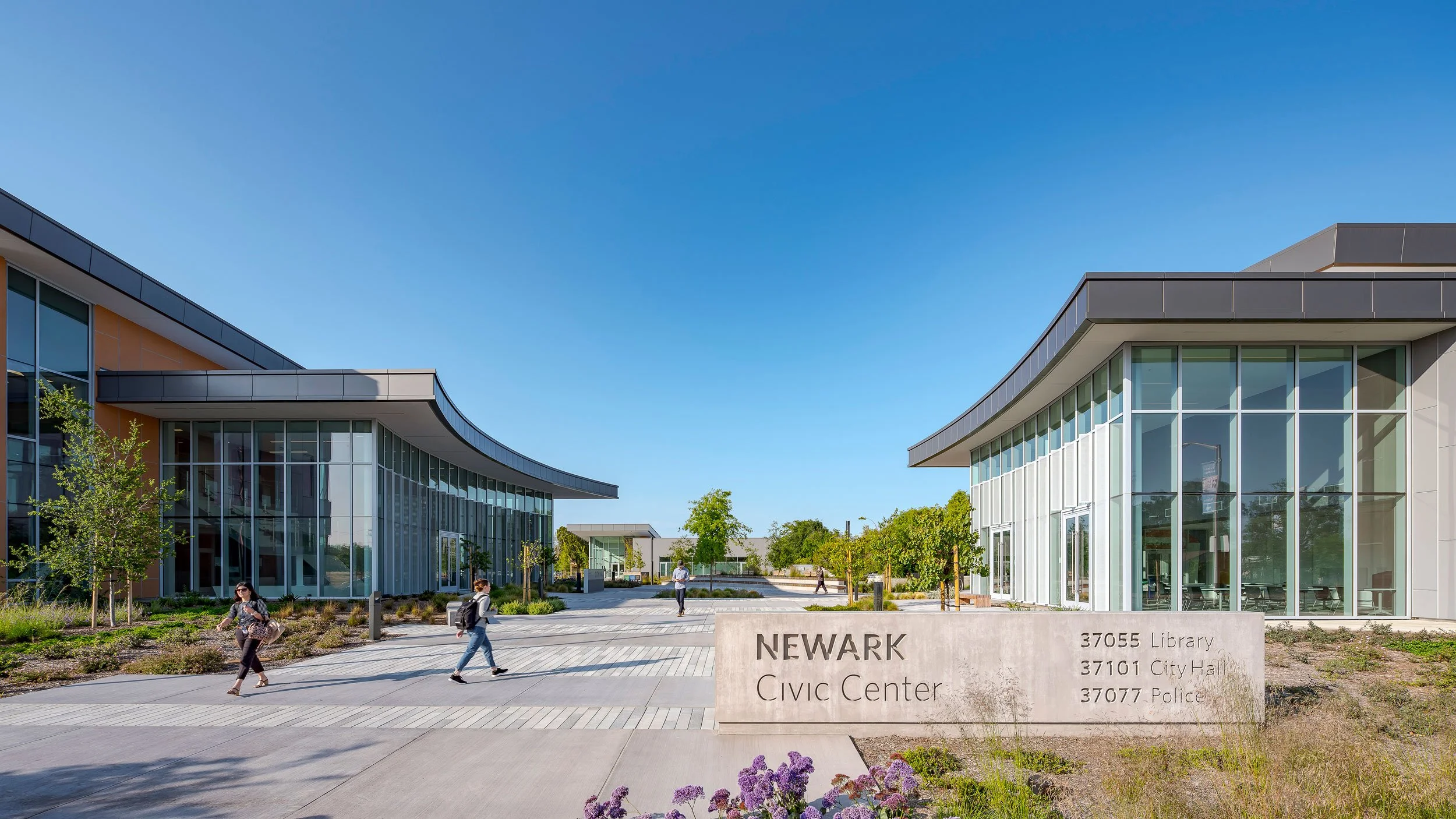 Exterior view of the Newark Civic Center with modern glass buildings, trees, and pedestrians on a sunny day.