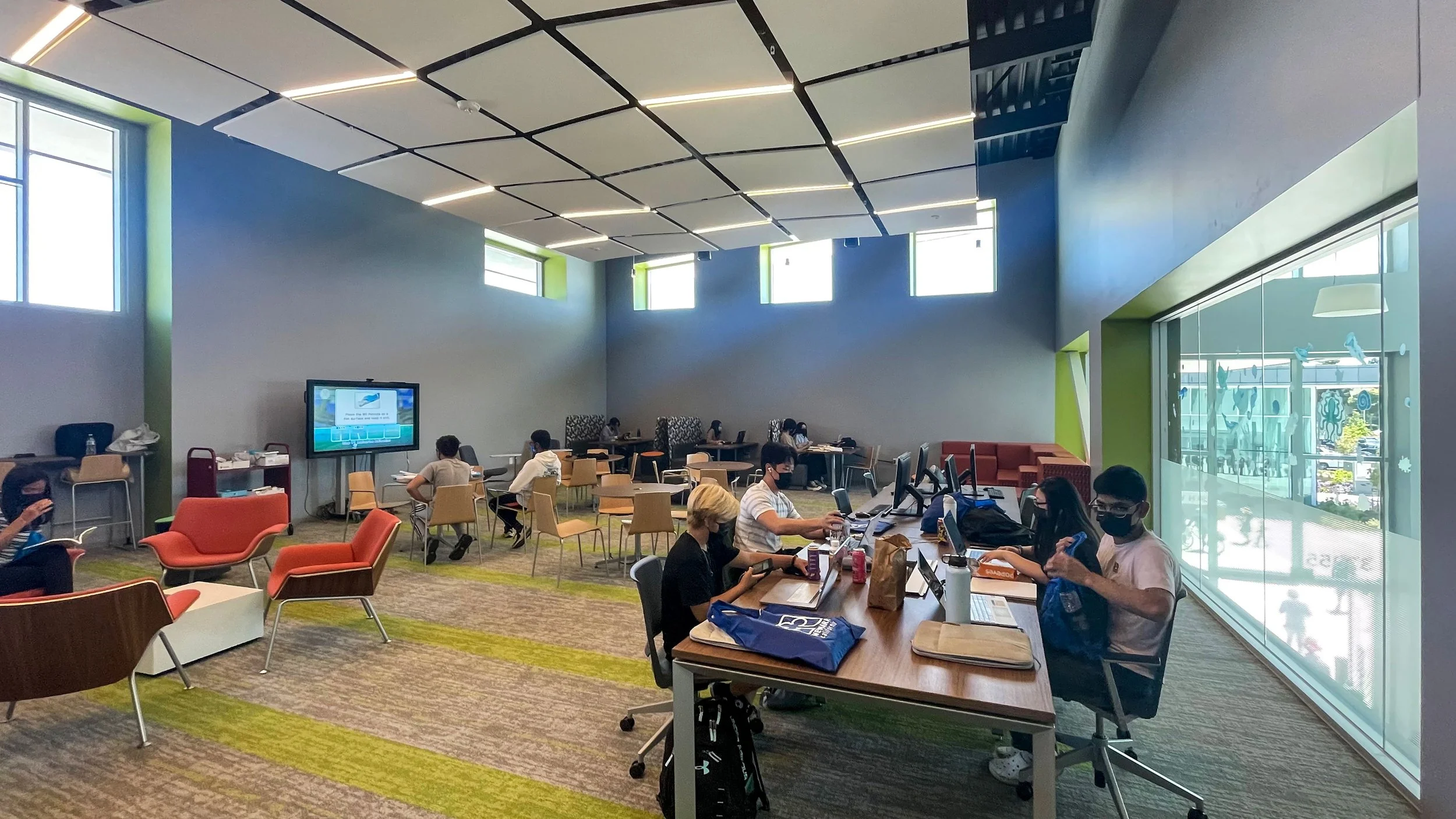 Indoor lounge area with people sitting at tables and working on computers, with large windows, modern ceiling design, and colorful chairs.