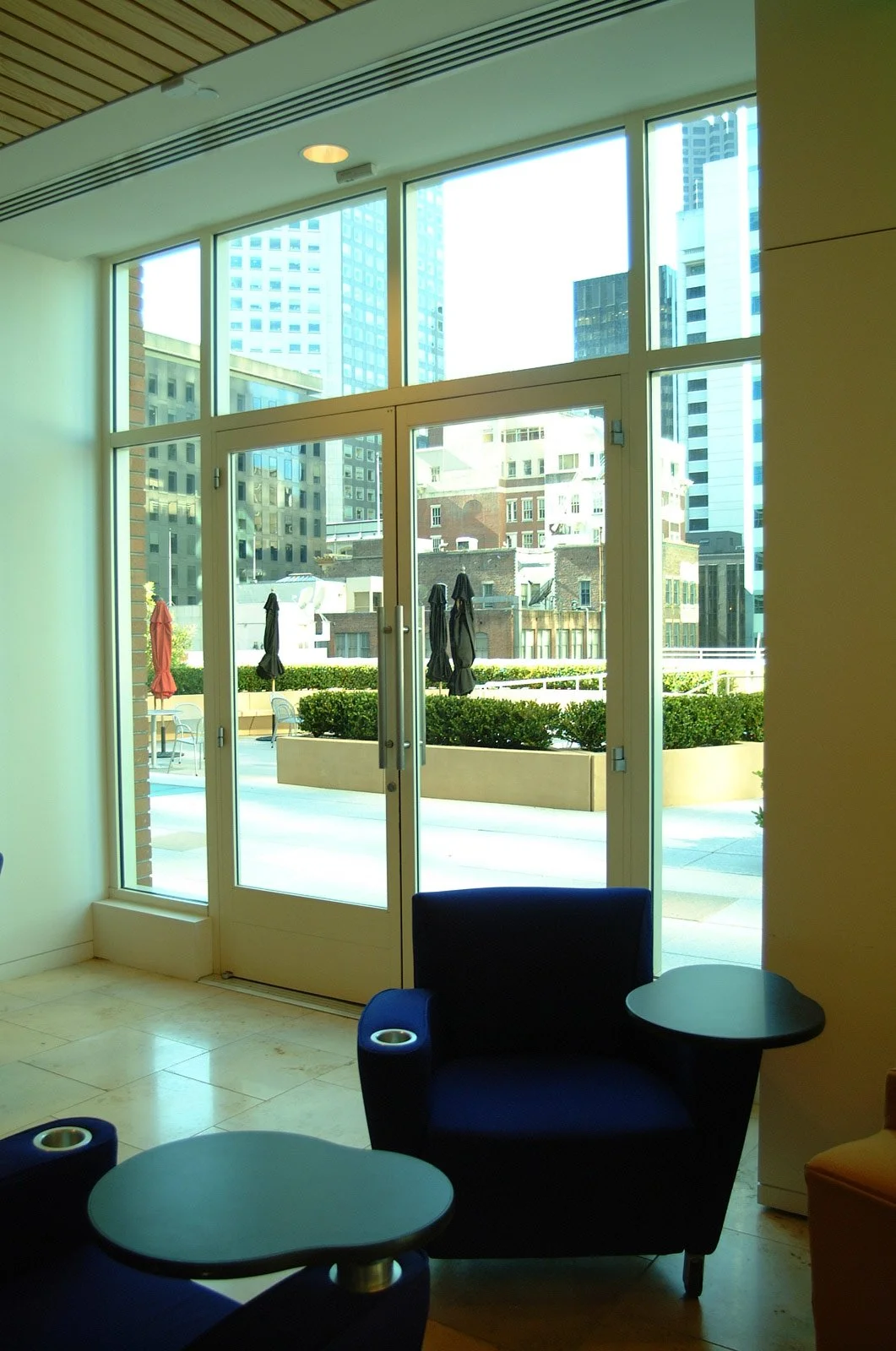 Interior of a modern lounge with large glass windows and door, overlooking a cityscape with tall buildings and an outdoor patio with closed umbrellas and seating.