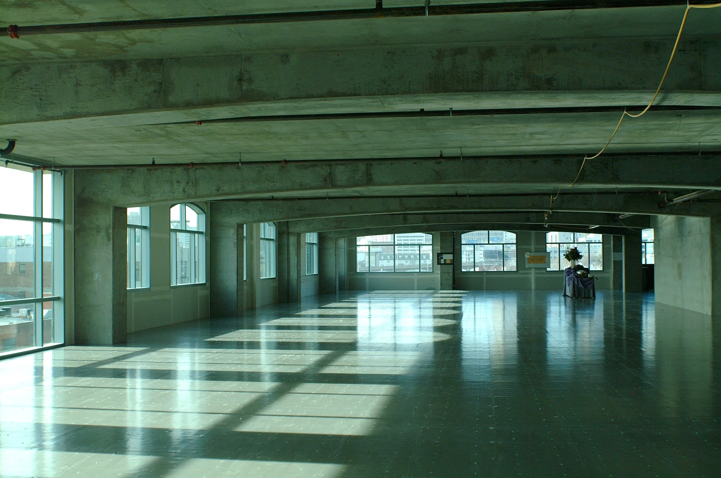 Empty industrial-style room with concrete ceiling and large windows letting in sunlight, casting shadows on the dark shiny floor.