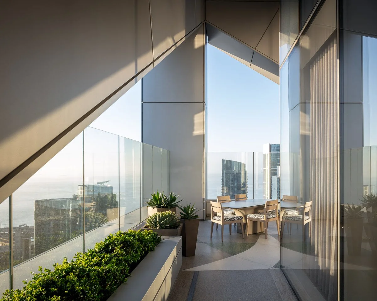 A modern high-rise balcony with glass railings, outdoor seating table with six chairs, and potted plants, overlooking a city skyline with tall buildings and a clear sky.