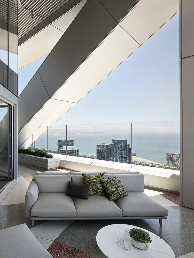 Modern living room with a white sofa, decorative pillows, and a round coffee table. Large windows and an open balcony overlook a city skyline and body of water.