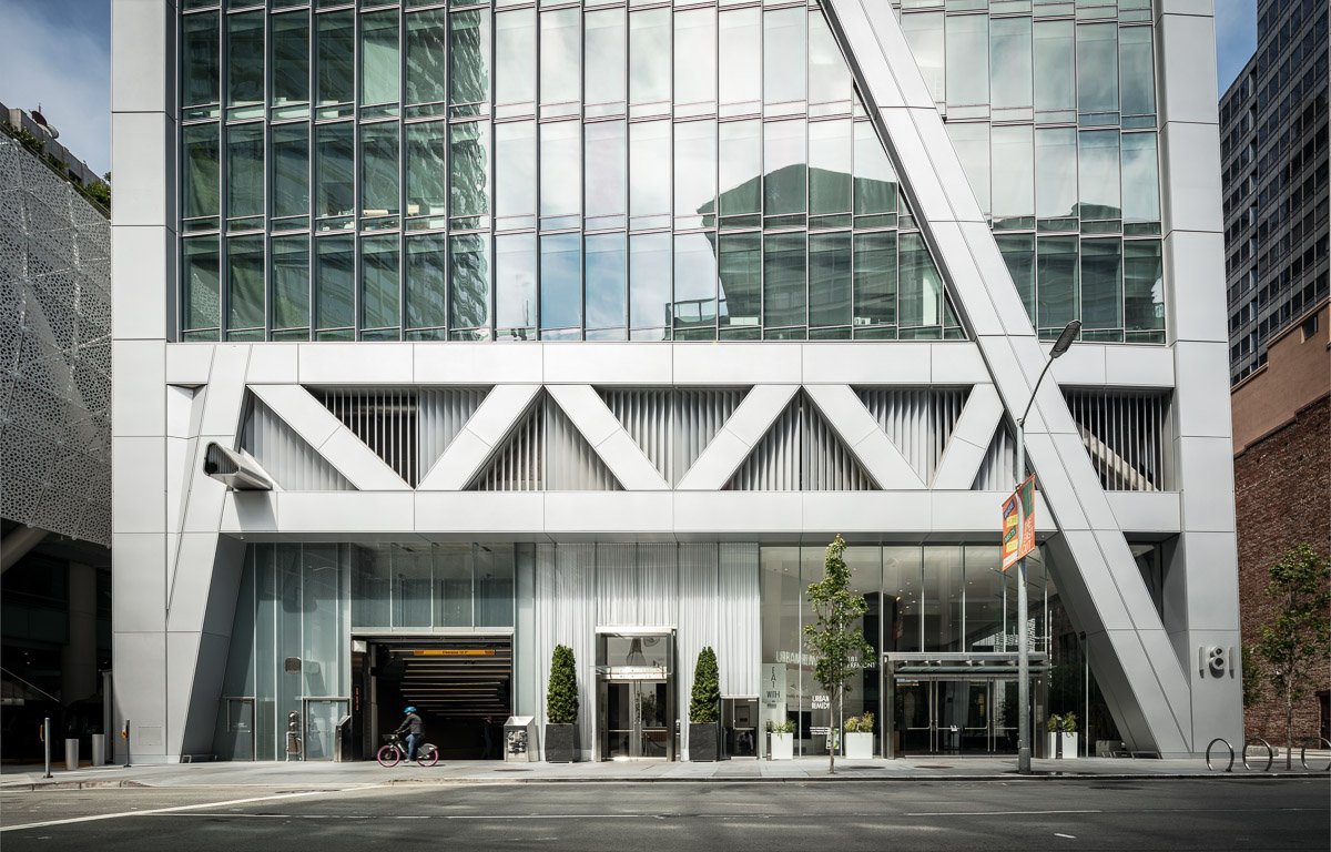 Modern multi-story building with glass windows and geometric metal architecture at the entrance, with a cyclist and a streetlamp in front.