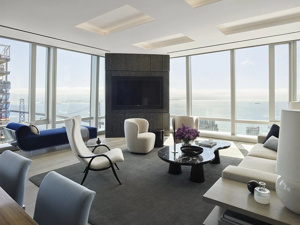Modern living room with large floor-to-ceiling windows overlooking a city and water, featuring contemporary furniture including a white sofa, white and blue chairs, a black marble coffee table with purple flowers, and a wall-mounted flat-screen TV.