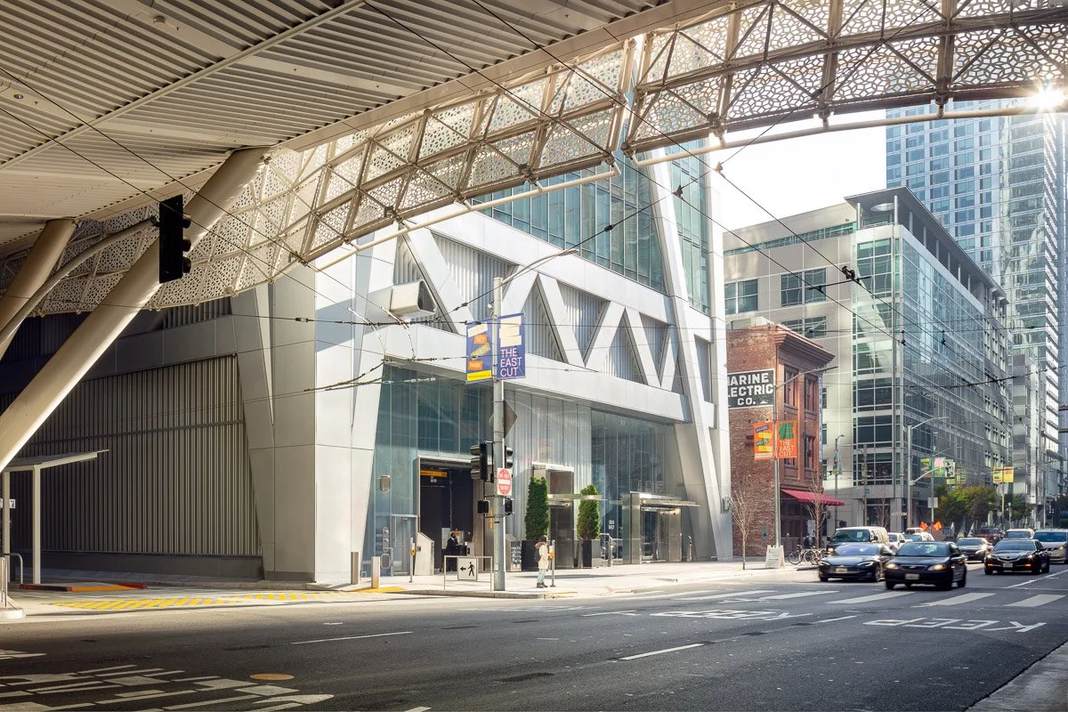 Modern building with large glass windows and architectural details, city street scene with cars and pedestrians, and a mix of older and newer buildings.
