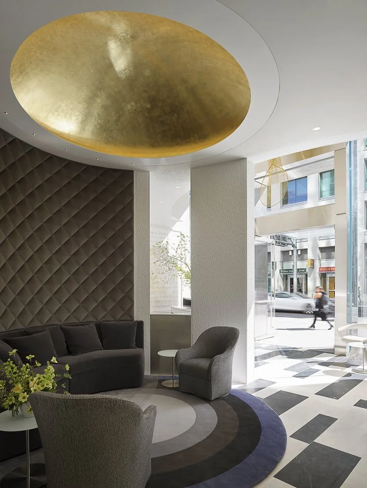 Modern hotel lobby with a gold ceiling feature, dark textured wall, black sofa, gray armchair, and a round patterned rug near large glass windows showing city street outside.