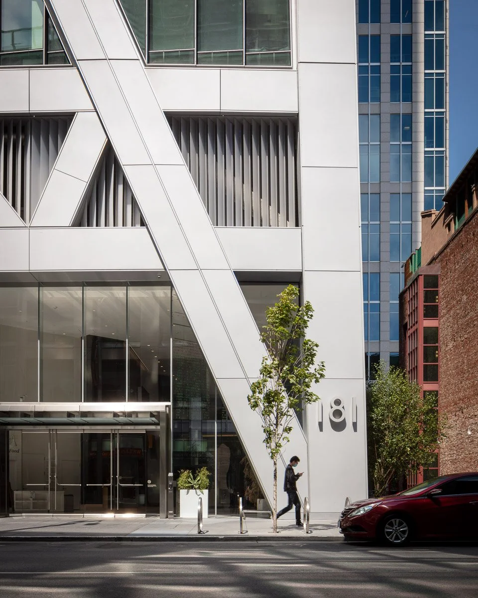 Modern white and glass building with diagonal architectural feature, trees, a person walking on sidewalk, and a red car parked on street.