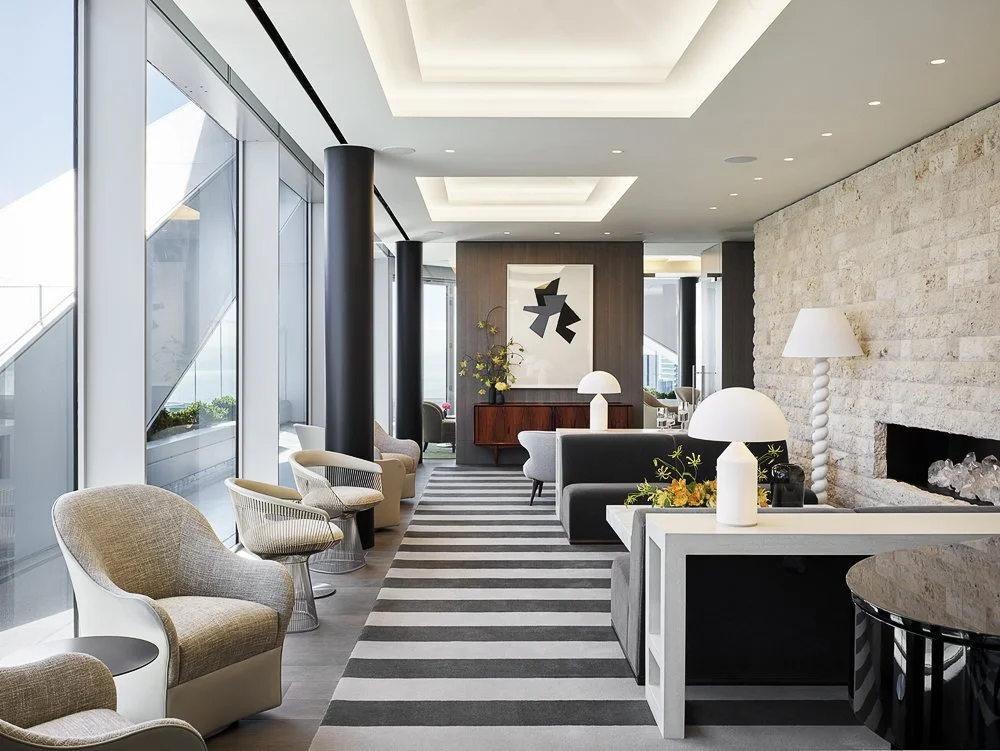 A modern, stylish living room with large glass windows, contemporary furniture, abstract artwork, pillows, lamps, and a stone fireplace.