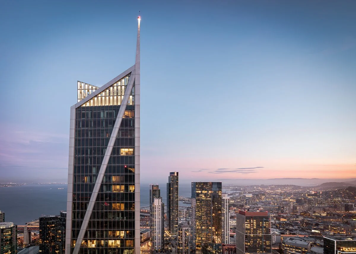 City skyline at dusk featuring a tall, modern skyscraper with distinctive slanted lines and glass windows, overlooking a cityscape with other tall buildings, water, and mountains in the distance.