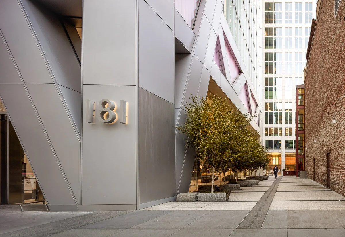 Urban alleyway with modern building on the left displaying the number "181" and an older brick building on the right. Paved sidewalk with trees in planters and a person walking in the distance.
