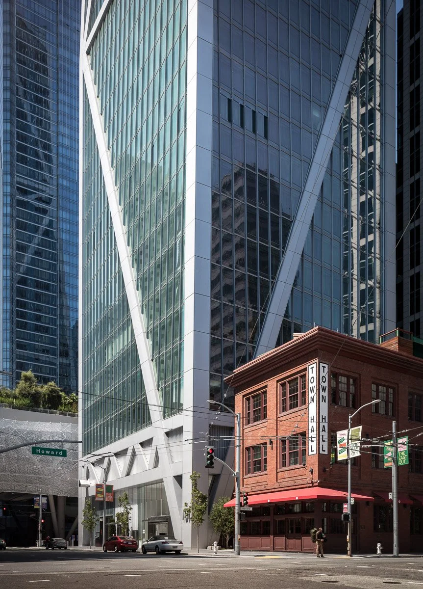 Tall modern glass skyscraper with steel beams and a smaller red brick building with a sign that says 'Town Hall' at street corner with cars and pedestrians.