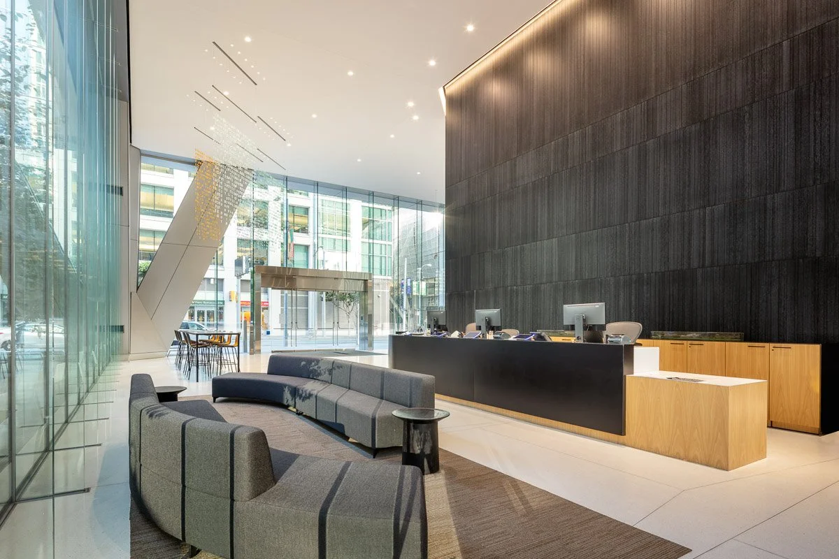 Modern hotel or office lobby with a sleek black reception desk and a gray curved sofa near the entrance, large glass windows, and wooden accents.