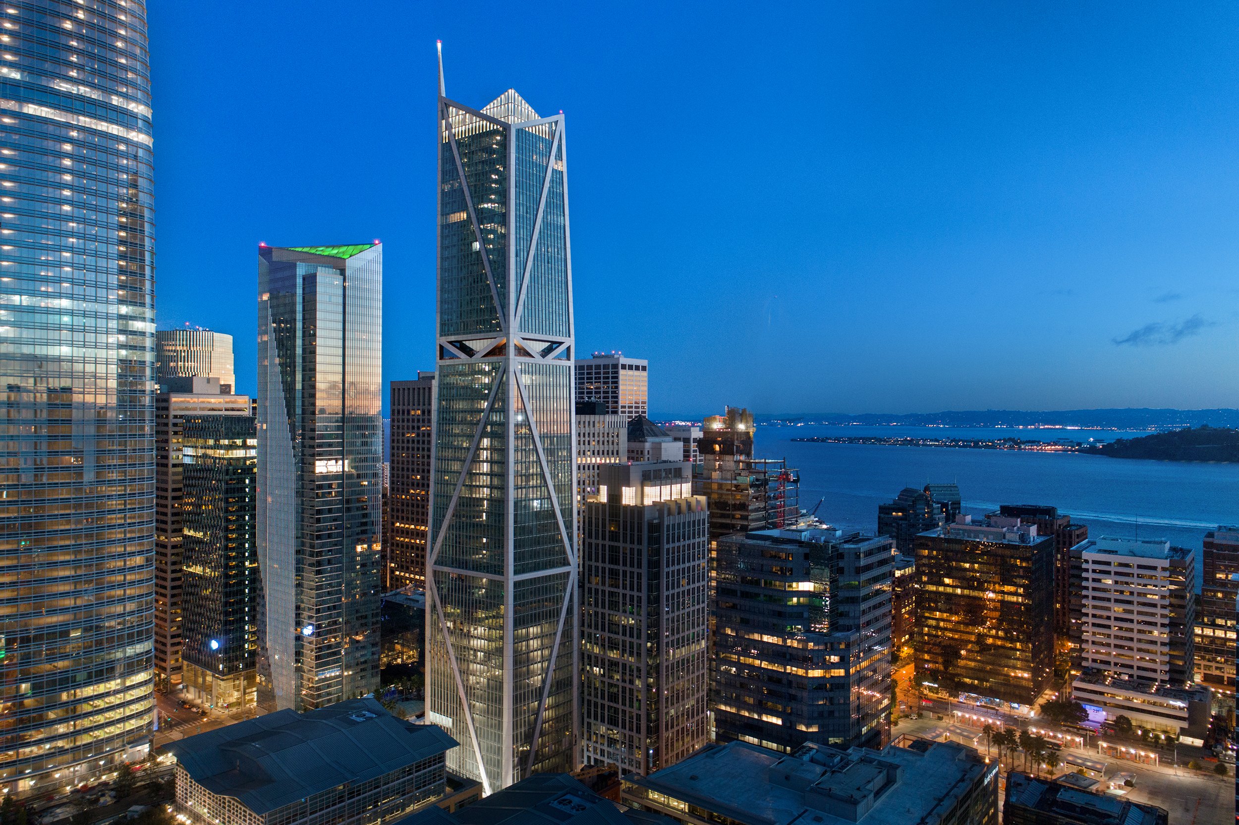 Aerial view of a city skyline at dusk, featuring tall modern skyscrapers along the waterfront with illuminated windows and a blue sky.