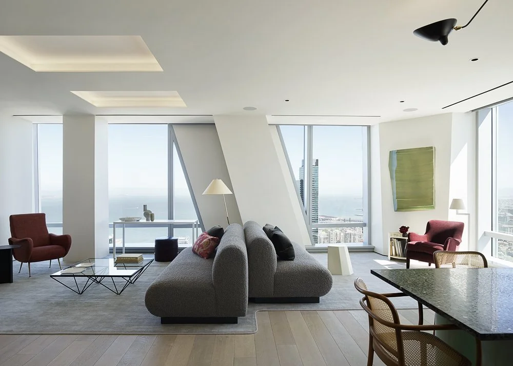 Modern living room with large windows showing city view, gray sofa, red armchairs, coffee table, artwork, and minimal decor.