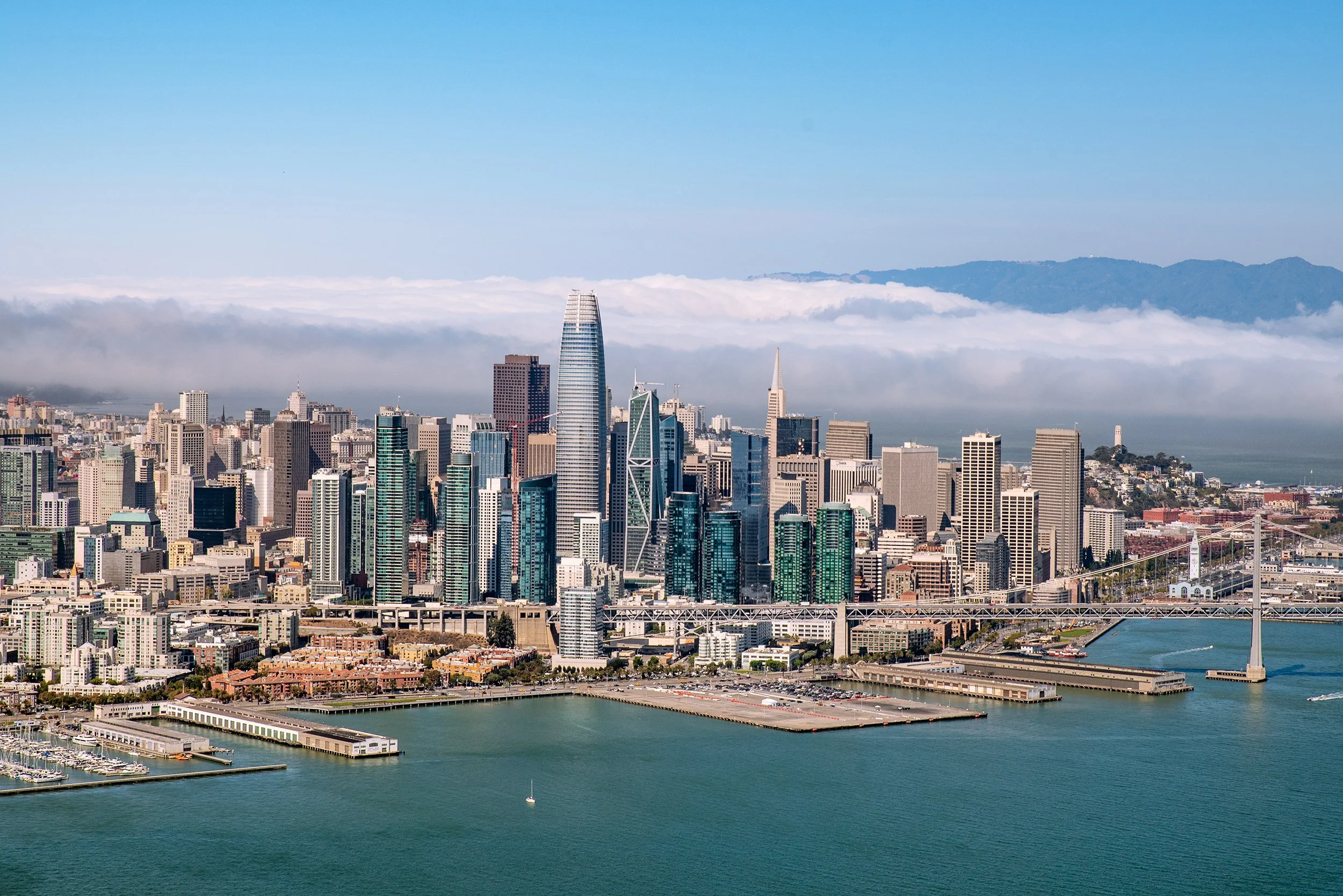 Aerial view of San Francisco skyline with tall skyscrapers, bay bridge, water, and mountain range in the background under blue sky.