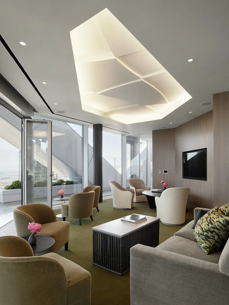 Modern hotel lounge with beige and white chairs, a gray sofa with patterned pillows, a black coffee table with books, pink flowers on side tables, large windows with city view, and a unique ceiling light fixture.