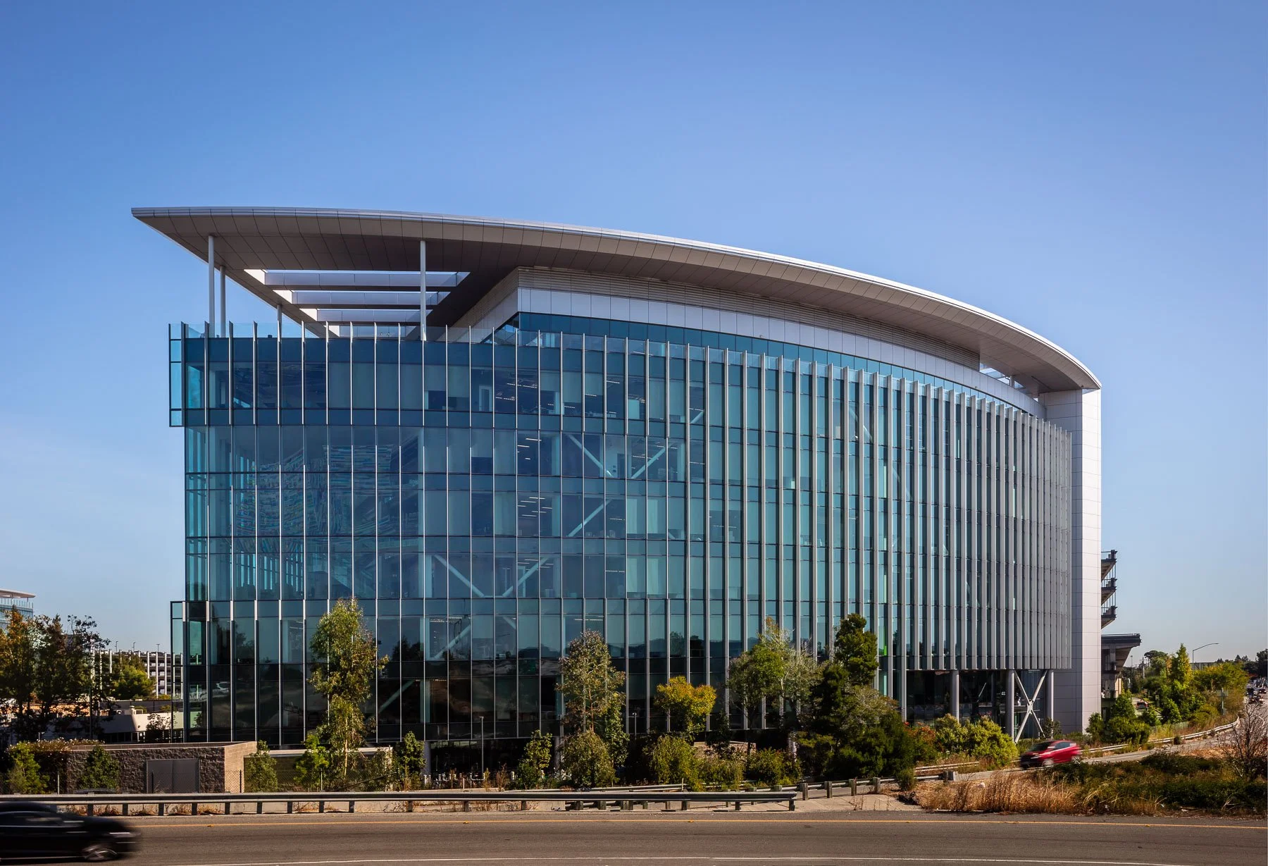 Modern office building with glass exterior, trees, and a highway in the foreground under a clear blue sky.