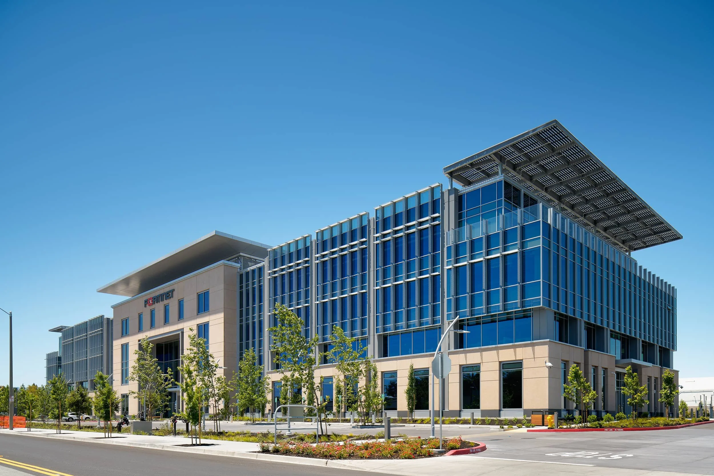 Fortinet Global Headquarter, Modern multi-story office building with blue glass windows and solar panels on the roof, surrounded by trees and a paved street.