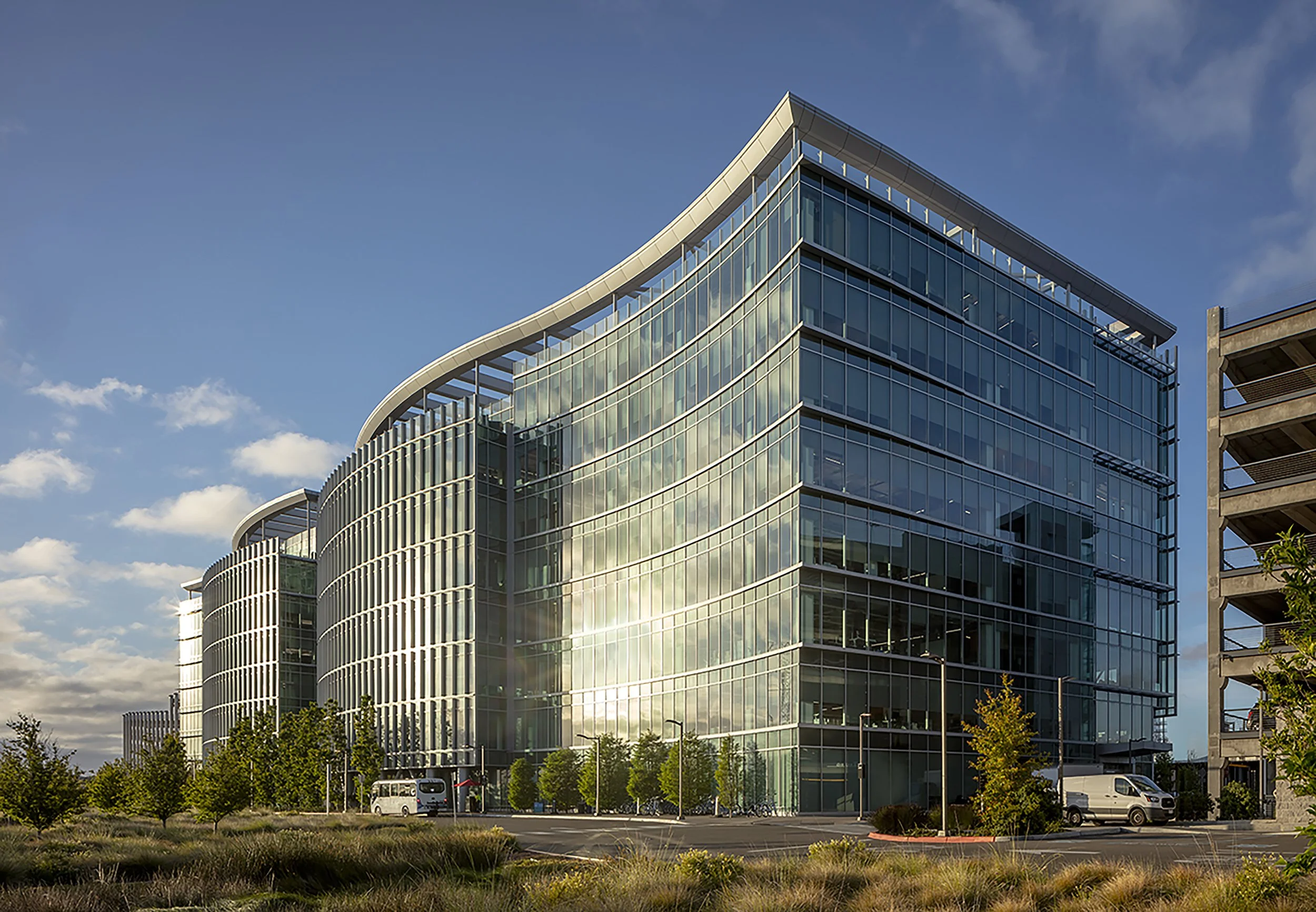 Modern glass office building reflecting the sky and clouds, with a curved design and surrounded by trees and parked vehicles.