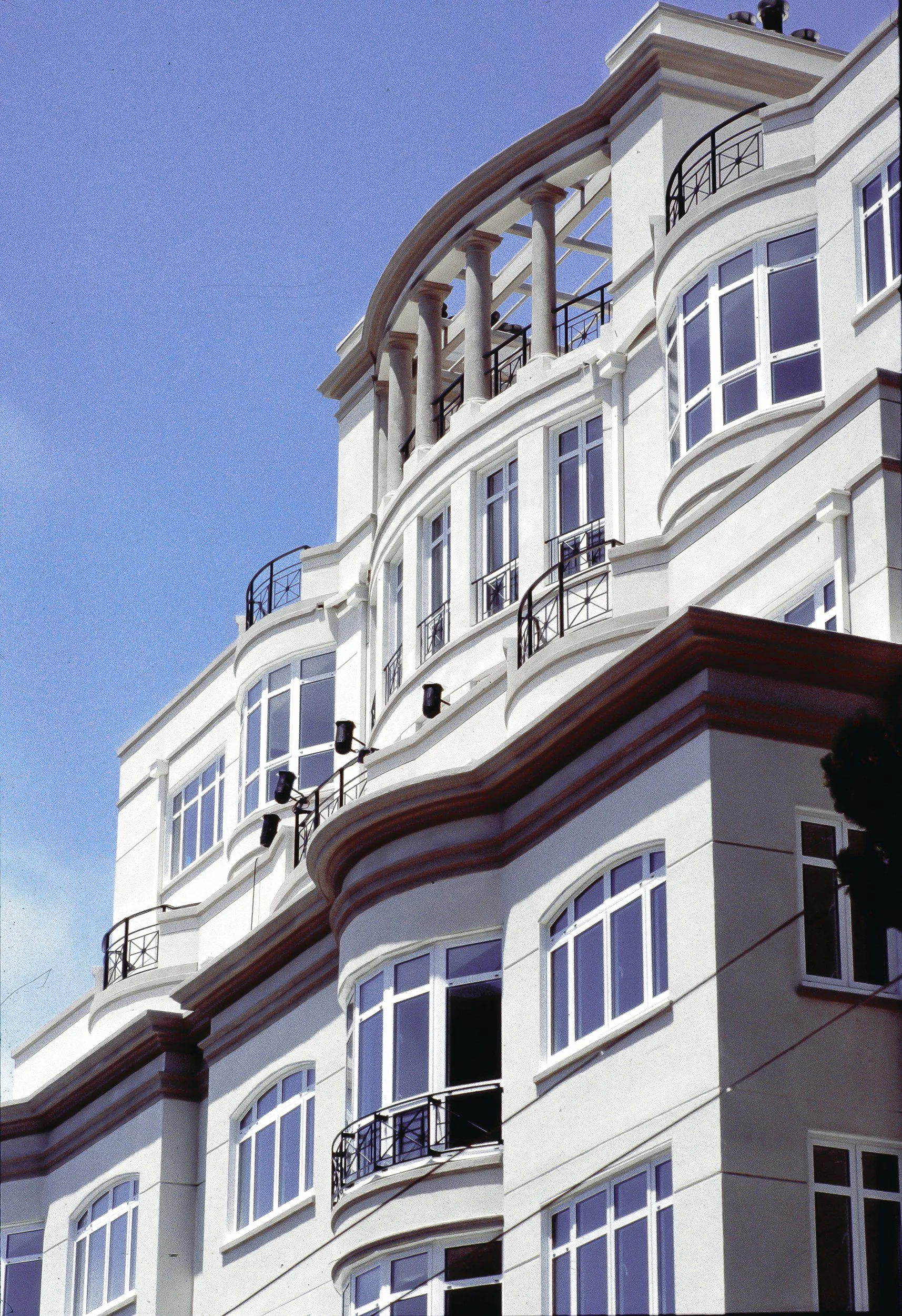 A multi-story modern building with white walls, curved balconies with black railings, large windows, and decorative architectural details, set against a clear blue sky.
