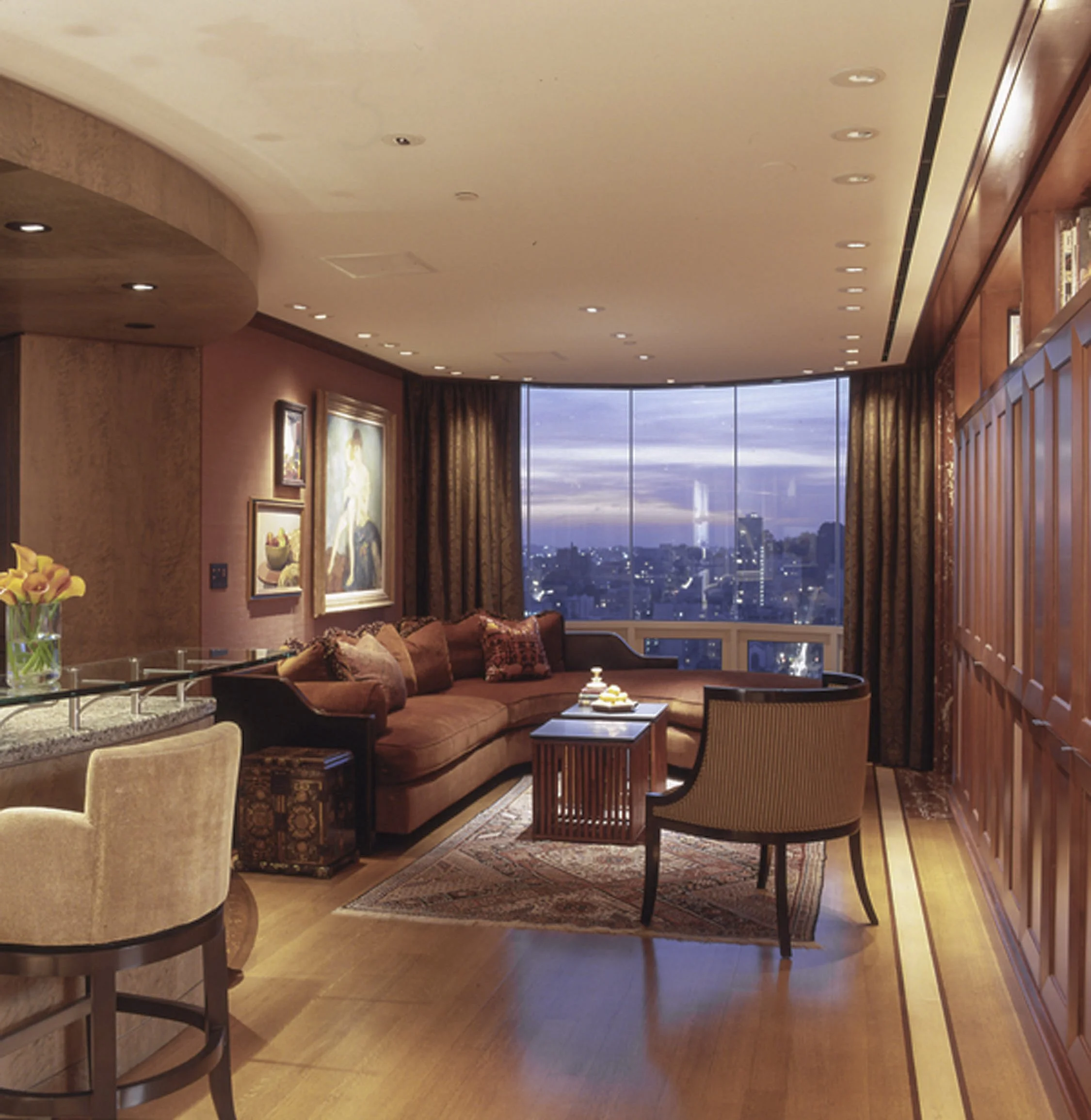 A luxurious living room with a large corner sofa, a single armchair, a coffee table, framed artwork on the walls, and large windows with city views at dusk.