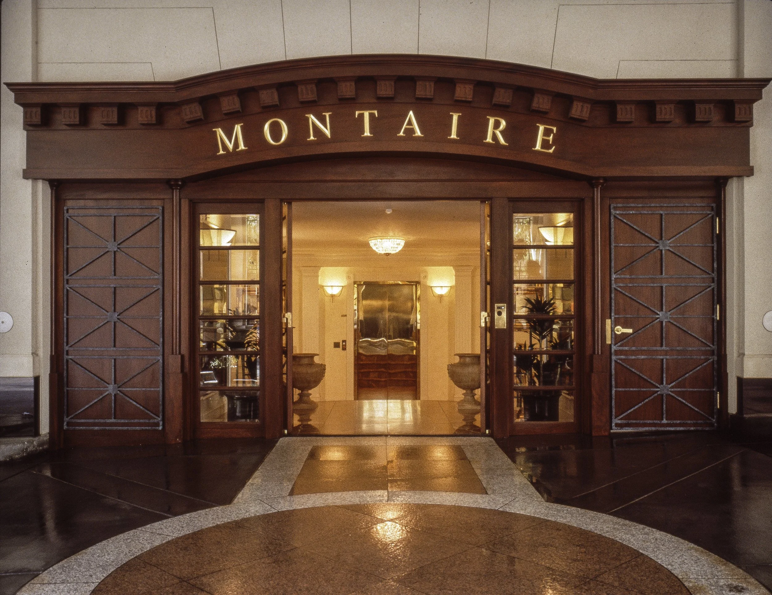 The entrance to a building with the sign 'Montaire' above the door, wooden doors partially open revealing a well-lit lobby with plants and decorative vases.