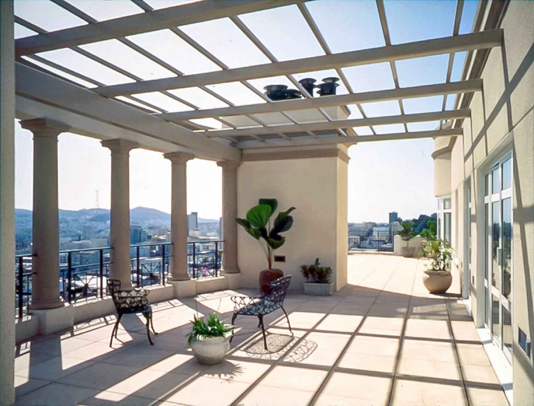 Sunlit rooftop terrace with potted plants, metal chairs, a pergola casting striped shadows, and cityscape in the background.