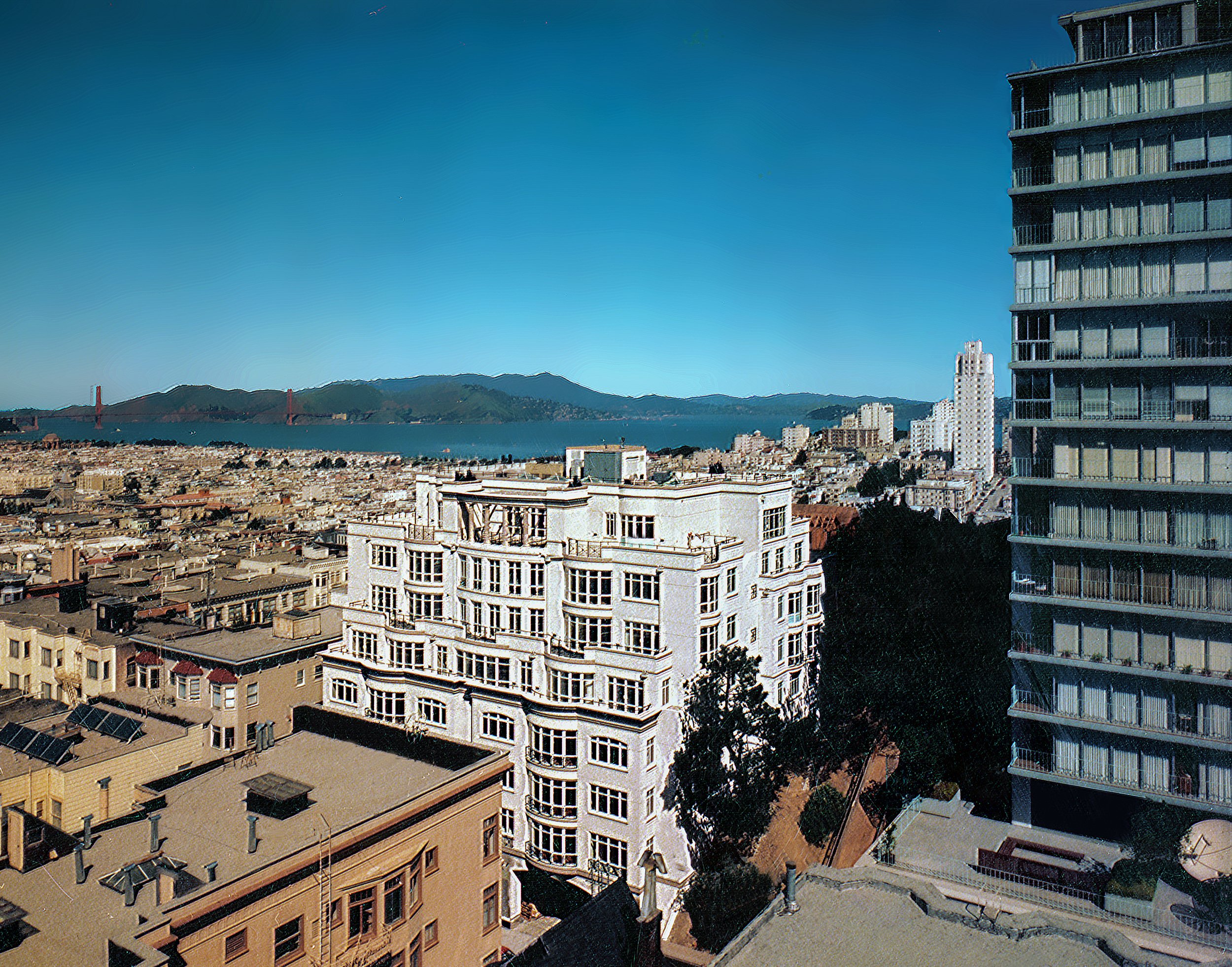 City skyline with a large white building in the foreground, other buildings around, the San Francisco Bay and hills in the background, and blue sky overhead.