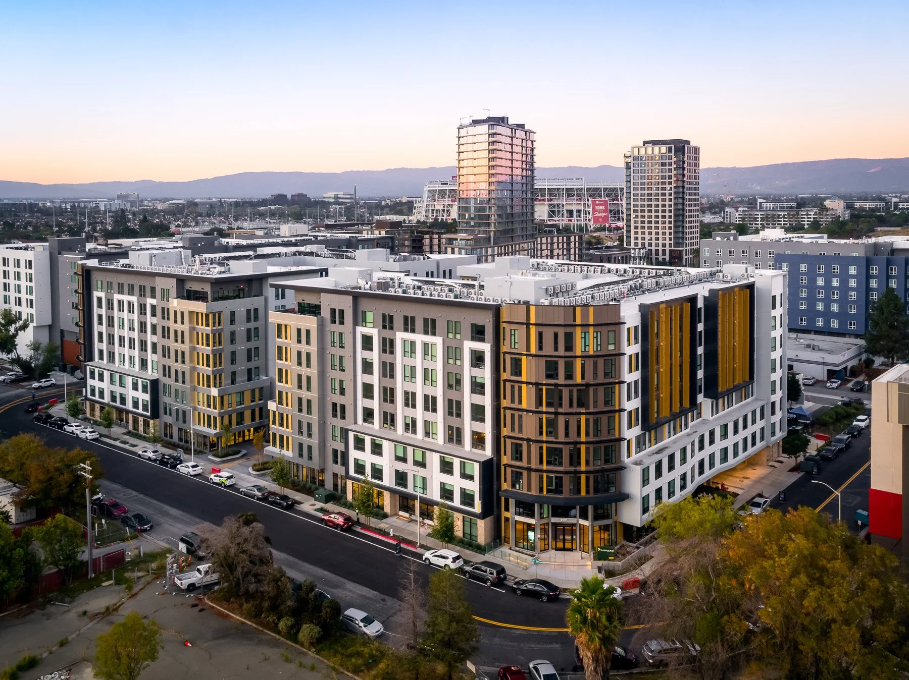 Modern multi-story apartment buildings on a city street at dusk, with parked cars and trees in the foreground.